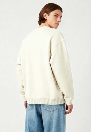 Felpa oversize beige chiaro con polsini e orlo a costine, realizzata in materiale morbido. Abbinata a jeans in denim blu chiaro.