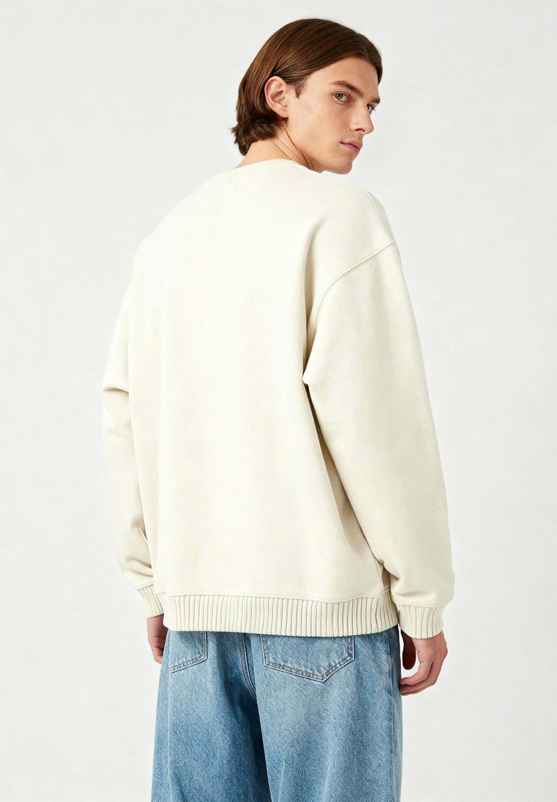 Felpa oversize beige chiaro con polsini e orlo a costine, realizzata in materiale morbido. Abbinata a jeans in denim blu chiaro.