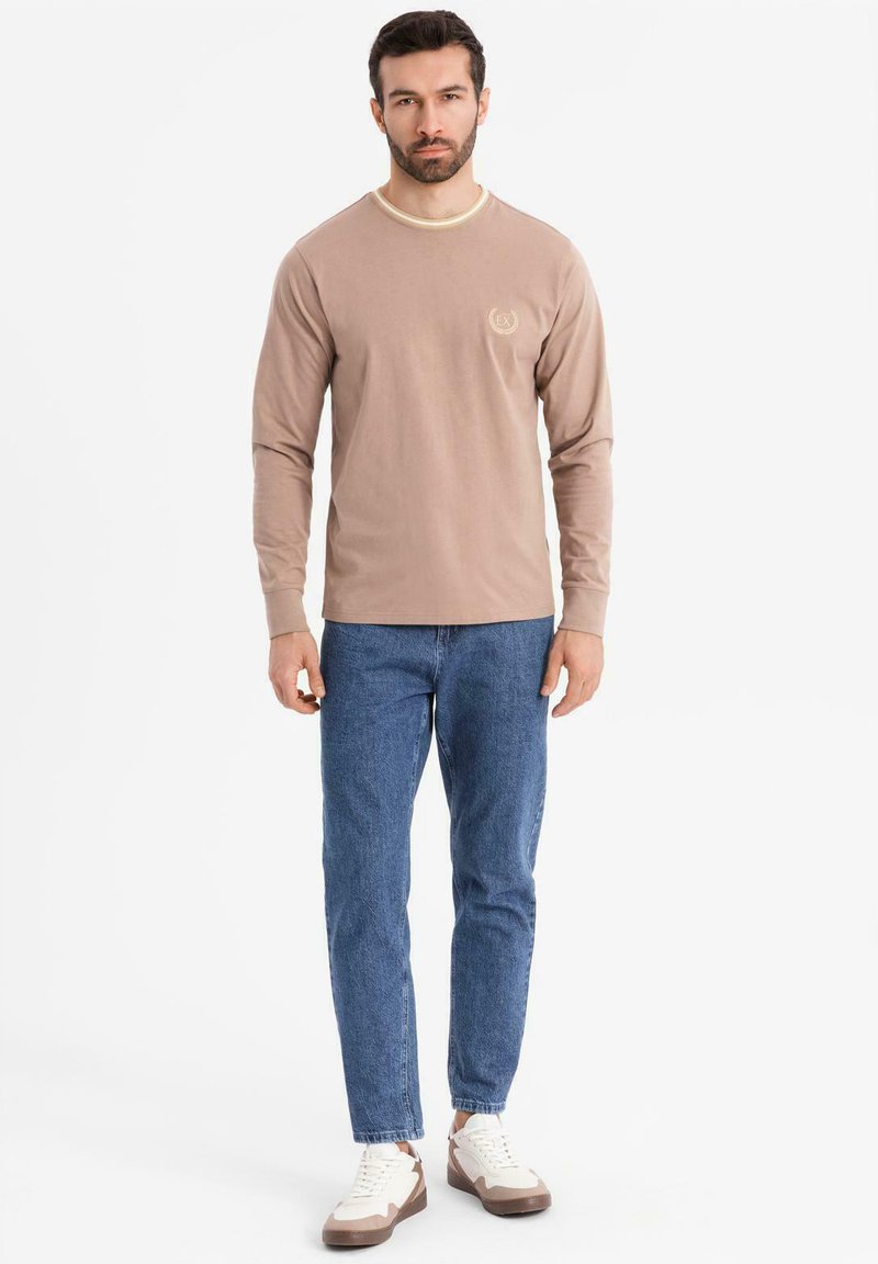 Uomo in piedi che indossa una camicia a maniche lunghe beige, jeans blu e sneakers bianche su uno sfondo bianco semplice.