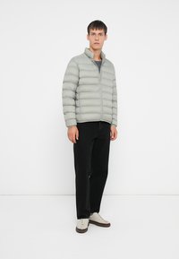 Lys grå polstret jakke med høy krage, glidelås og horisontale sømmer, kombinert med sorte bukser og beige sneakers.