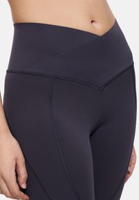 Leggings noirs taille haute avec un design de ceinture croisée, texture lisse et détail de surpiqûres contrastantes le long des coutures.
