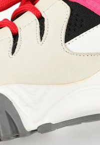 Gros plan sur le côté d'une basket montrant des panneaux blancs et noirs, des lacets rouges et des détails texturés sur la semelle grise et blanche.