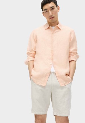Mann trägt ein hell pfirsichfarbenes, geknöpftes Hemd über einem weißen T-Shirt und beigen Shorts, steht mit den Händen in den Taschen vor einem einfarbigen Hintergrund.