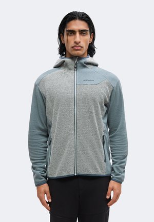 Jonge man met donker haar, gekleed in een grijze zip-up hoodie met gestructureerde frontpanelen en zwarte broek, kijkt recht vooruit tegen een effen achtergrond.