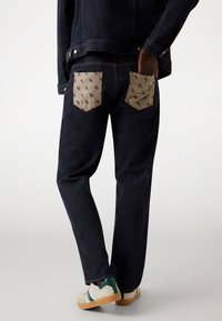 Dark blue denim jeans with tan patterned patch pockets, straight cut, and minimal stitching details. Paired with white and green sneakers.