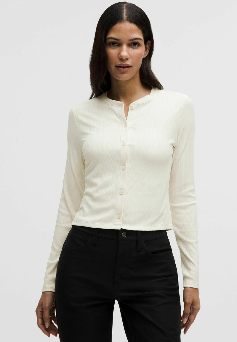 Frau trägt ein eng anliegendes cremefarbenes, geripptes, langärmeliges, geknöpftes Oberteil und hoch taillierte schwarze Hosen vor einem einfarbigen Hintergrund.