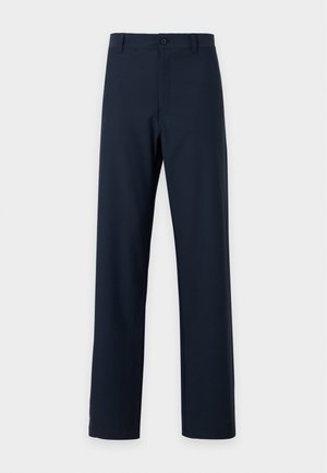 Jeune homme debout, les mains dans les poches, portant un t-shirt blanc à manches courtes, un pantalon foncé et des baskets blanches sur un fond uni.