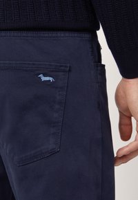 Pantaloni blu navy con una tasca posteriore decorata da un ricamo di un bassotto azzurro. Texture liscia, design aderente e tasche laterali per le mani visibili.