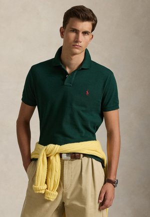 Νέος άνδρας που φοράει σκούρο πράσινο πόλο, μπεζ παντελόνια, κίτρινο πουλόβερ δεμένο γύρω από τη μέση και ρολόι χειρός, στέκεται μπροστά σε απλό φόντο.