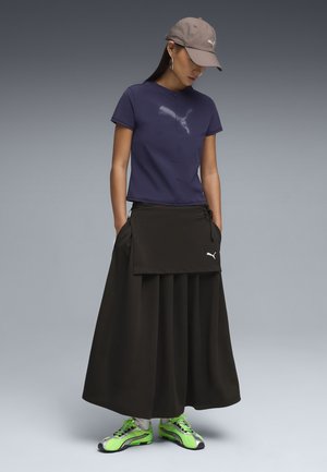 Donna con maglietta Puma blu navy, gonna lunga nera con borsa attaccata, cappellino beige Puma e sneaker verde neon, in piedi con le mani nelle tasche.