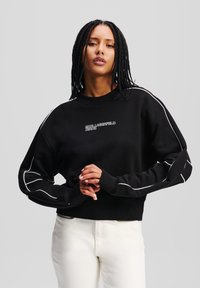 Čierna mikina vyrobená z bavlny, s rebrovaným okrúhlym výstrihom, bielym lemovaním pozdĺž rukávov a logom "KARL LAGERFELD JEANS" na hrudi.