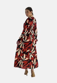 Lange, zwarte jurk met een levendig rood en crèmekleurig bloemenpatroon. Heeft lange mouwen en een getailleerde taille, gecombineerd met beige schoenen.