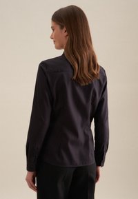 Blusa blu navy con maniche lunghe, colletto a punta e polsini con bottone. Presenta una texture liscia e un dettaglio nella parte posteriore con yoke, abbinata a pantaloni sartoriali.