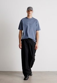 Siva pamučna majica s kratkim rukavima, koja sadrži suptilan logotip, kombinirana s labavim crnim hlačama i crnim cipelama. Model nosi sivi šešir.