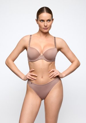 Femme portant un soutien-gorge et une culotte marron clair assortis, debout les mains sur les hanches sur fond blanc.