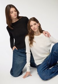 Due modelli indossano maglioni in maglia leggera: uno nero, l'altro bianco. Entrambi abbinati a jeans larghi e sbiaditi di colore blu.