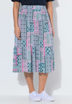 Kobieta w niebiesko-różowo-zielonej wzorzystej spódnicy o długości do połowy łydki, w białych trampkach, stojąca na tle gładkiej, białej ściany.