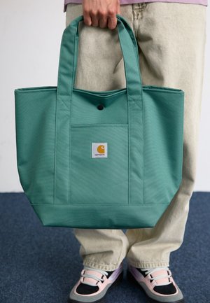 Persona que sostiene una gran bolsa verde Carhartt por las asas, con pantalones beige y zapatillas rosas y negras.