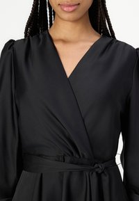 Robe noire à manches longues avec un design croisé, présentant un décolleté en V profond, des épaules froncées et une ceinture nouée à la taille, fabriquée en tissu lisse.