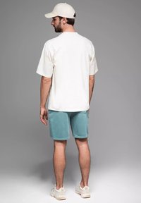 T-shirt blanc à manches courtes, short bleu clair et baskets blanches. Casquette neutre portée à l'envers. Tenue décontractée avec une coupe ample.