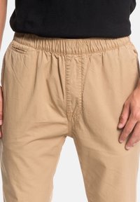 Beiga elastiska midjebyxor gjorda av lättviktsmaterial, med en slät yta, praktiska sömmar och sidofickor för bekvämlighet.