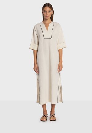 Robe en lin de couleur blanc cassé avec un col en V et des passepoils noirs. Elle est dotée de manches courtes et d'ourlets fendus sur les côtés pour une plus grande liberté de mouvement. Des sandales plates complètent le look.