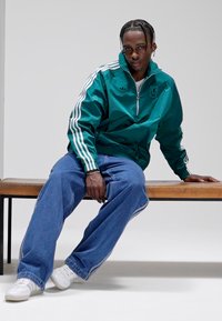 Grönblå dragkedjejacka med vita ränder, kombinerad med blå vida jeans och vita sneakers, sittande på en brun bänk mot en vit bakgrund.