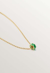 Collar de cadena de oro con un colgante de gema verde redonda y acentos florales, descansando sobre un fondo claro.