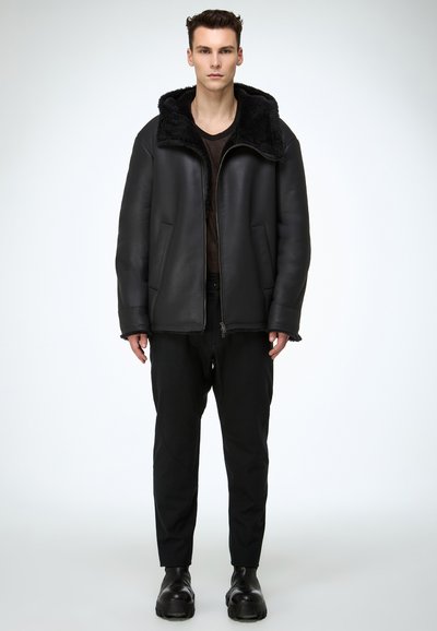 Mann steht und trägt eine schwarze Lammfelljacke mit großem Kragen, schwarze Hose, schwarze Stiefel und ein dunkles Hemd vor einem einfarbigen weißen Hintergrund.