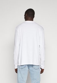 Mand vendt væk iført en ensfarvet hvid langærmet skjorte og lyseblå cowboybukser mod en neutral baggrund.