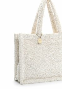 Kermanvärinen tekstituotteinen olkalaukku, jossa on lyhyet kahvat ja pieni pyöreä kullanvärinen riipus lähellä yläreunaa.