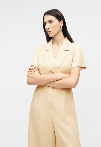 Γυναίκα με μακριά σκούρα μαλλιά που φοράει μπεζ ολόσωμη φόρμα σε στυλ σακακιού με κοντά μανίκια, τα χέρια σταυρωμένα, κοιτάζει προς το πλάι μπροστά σε λευκό φόντο.