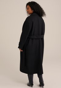 Černý vlněný kabát s páskem, oversized design, boční rozparky a manžety na knoflíky. Tkanina působí hladkým a strukturovaným dojmem.
