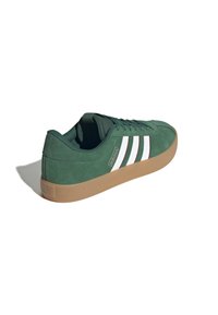 Zielony zamszowy sneaker Adidas z białymi paskami, gumową podeszwą i sznurowadłami, widok z boku i lekko zza pleców.