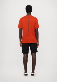 Tricou din bumbac roșu cu mâneci scurte, având pe spate un text vertical negru "EA7 EMPORIO ARMANI". Purta cu pantaloni scurți negri și adidași albi.