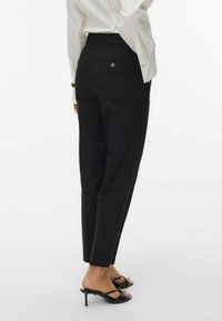 Mujer vestida con pantalones negros de sastre hasta los tobillos, blusa blanca y sandalias de tacón abiertas negras, de pie con una ligera inclinación.