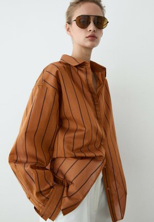 Kvinde iført oversized brun skjorte med sorte striber og hvide bukser, accessoriseret med store brune solbriller med tonede glas, mod en ensfarvet baggrund.