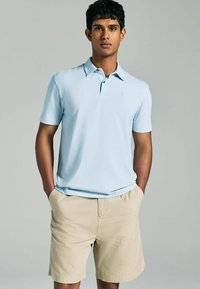 Hellblaues Poloshirt mit kurzen Ärmeln und kleinem Logo, kombiniert mit khakifarbenen Shorts. Der Stoff wirkt glatt und leicht.