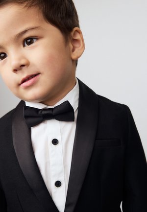 Ragazzo giovane che indossa una giacca da smoking nera, camicia bianca e papillon nero, guarda leggermente di lato con uno sfondo semplice.