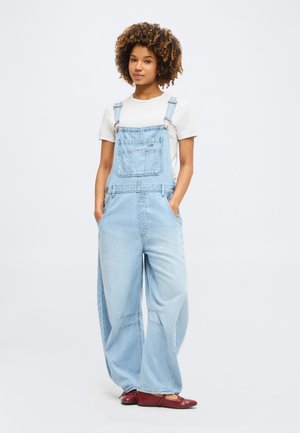 Νέα γυναίκα με σγουρά μαλλιά που φοράει ανοιχτό μπλε τζιν ολόσωμη φόρμα, λευκό κοντομάνικο πουκάμισο και κόκκινα επίπεδα παπούτσια, στέκεται με τα χέρια στις τσέπες.