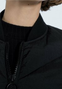 Zwarte bomberjack met een ritssluiting aan de voorkant, een geribde kraag en een gestructureerde stof. Close-up weergave die de stiksels en het ritssluitingsdetail benadrukt.