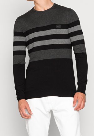 Mężczyzna ubrany w długi sweter w paski w odcieniach ciemnoszarym, czarnym i jasnoszarym, z białymi spodniami na jednolitym tle.