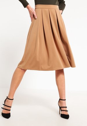 Faltenrock in Beige aus glattem Stoff, knielang mit einem taillierten Bund. Kombiniert mit schwarzen Riemchen-Pumps.