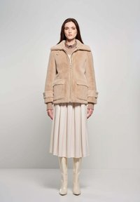 Beige faux fur jas met grote kraag en voorzakken, gecombineerd met een crèmekleurige geplooide rok en glanzende kniehoogte laarzen.