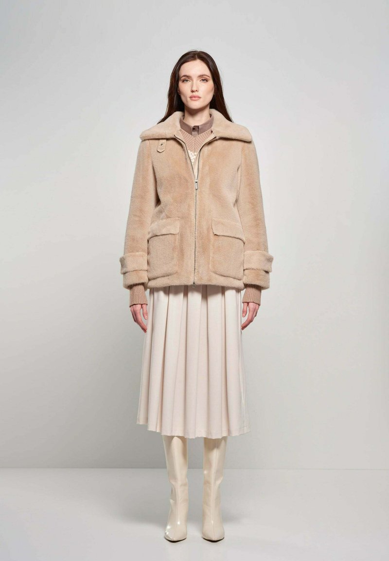 Beige faux fur jas met grote kraag en voorzakken, gecombineerd met een crèmekleurige geplooide rok en glanzende kniehoogte laarzen.