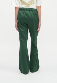 Cilvēks uzvilcis tumši zaļu platu bikses un baltu īsām piedurknēm kreklu ar dzeltenu grafiku, skatīts no aizmugures uz baltā fona.