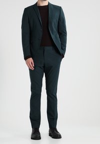 Mężczyzna w ciemnoniebieskim garniturze, czarnej bluzie i czarnych butach formalnych, stojący z jedną ręką w kieszeni na jednolitym tle.