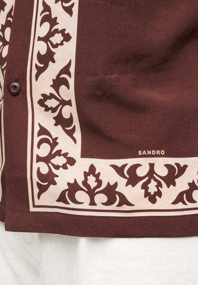 Καφέ τελείωμα πουκαμίσου με κρεμ φλοράλ σχέδιο περιγράμματος και σκοτεινό κουμπί, που δείχνει μερικό λογότυπο της μάρκας «SANDRO» κοντά στο κάτω άκρο.