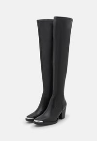 Botas de cuero negras hasta la rodilla con tacón cuadrado, puntera redondeada y detalles plateados en la puntera. Textura suave con costuras visibles y detalle de cremallera.