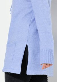 Hellvioletter Strickpullover mit seitlichem Schlitz, gerippten Bündchen und einer Fronttasche. Weiche Textur und lockere Passform, seitlich gezeigt.